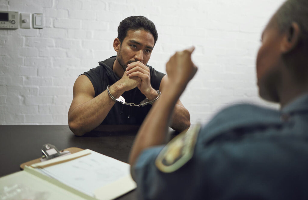 A man in handcuffs sits at a table while being questioned by a law enforcement officer, symbolizing individuals facing prescription drug fraud charges in Binghamton.