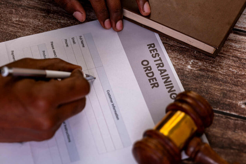 Person signing a 'RESTRAINING ORDER' document with a gavel and legal book nearby, symbolizing legal action.