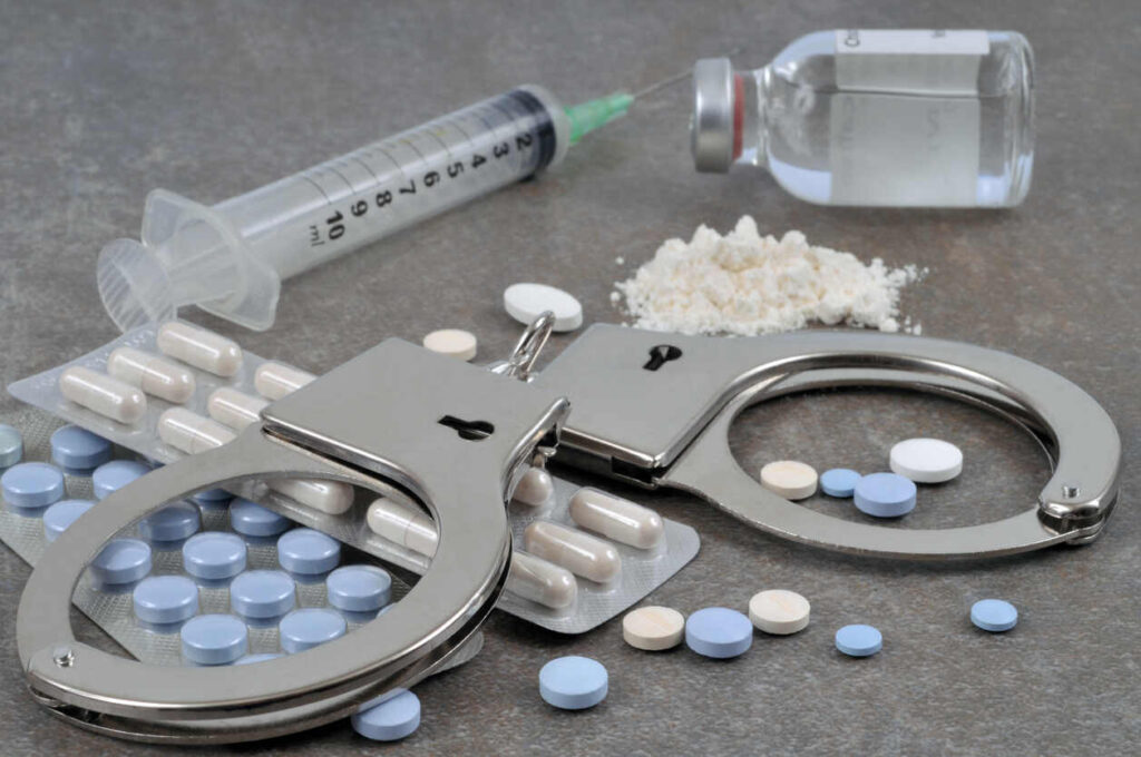 Handcuffs, blister packs of pills, syringes, and a vial on a gray surface, symbolizing drug-related charges and search warrants.