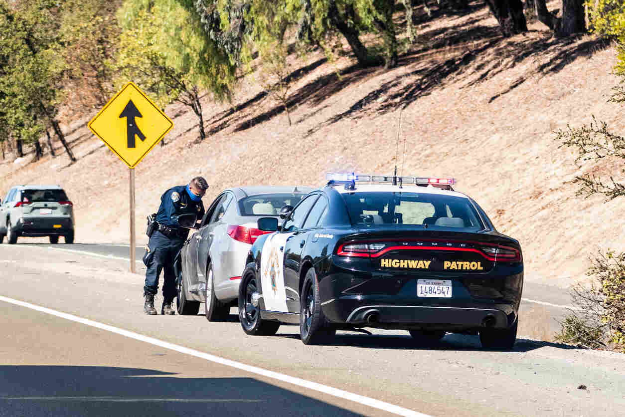 Police officer conducting a traffic stop on a highway with a highway patrol car and a silver vehicle, with a yellow road sign indicating a merge ahead.