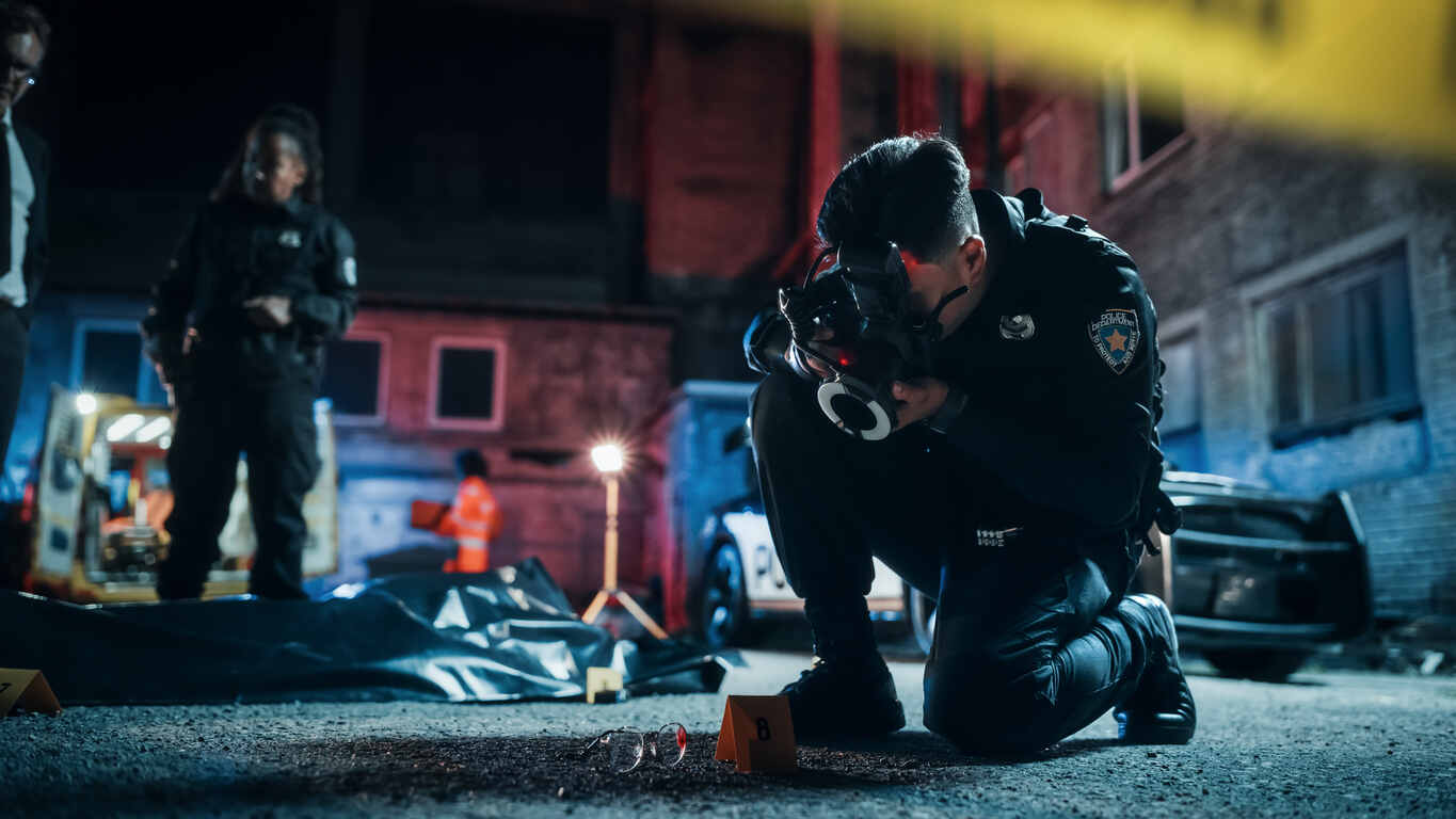 Police officer photographing evidence at a crime scene with yellow markers, while other officers and emergency responders are in the background, indicating an investigation in a criminal case.