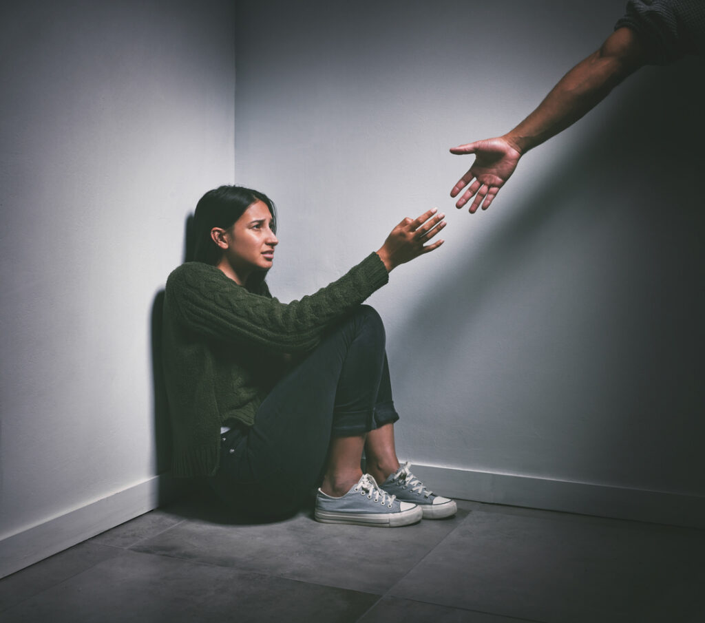 Shot of a young woman sitting in the corner of a dark room with a hand reaching out to help her with facing Sex Crime